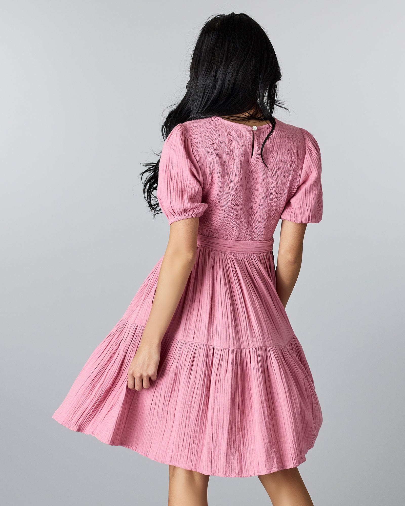 Woman in a pink, short sleeved, knee-length dress with smocking on bodice and a tie at the waist.