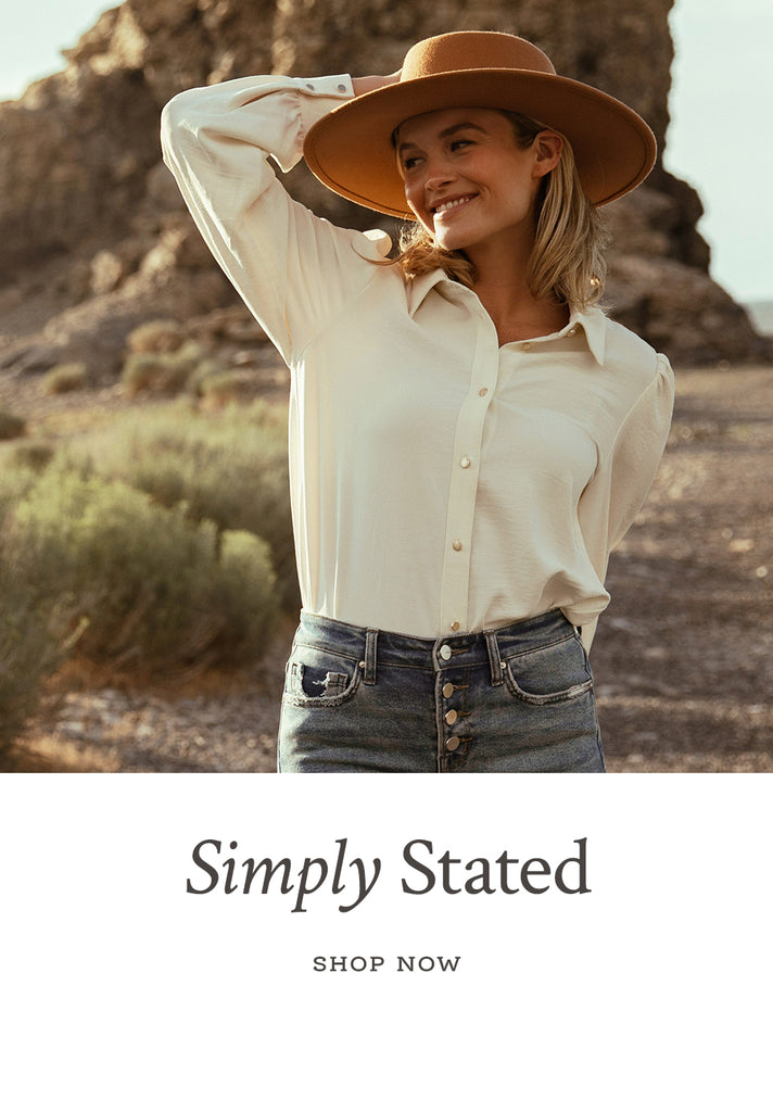 Text over image "Simply Stated - Shop Now". The image is of a women at the beach wearing a white button shirt, blue jeans, and a tan hat.