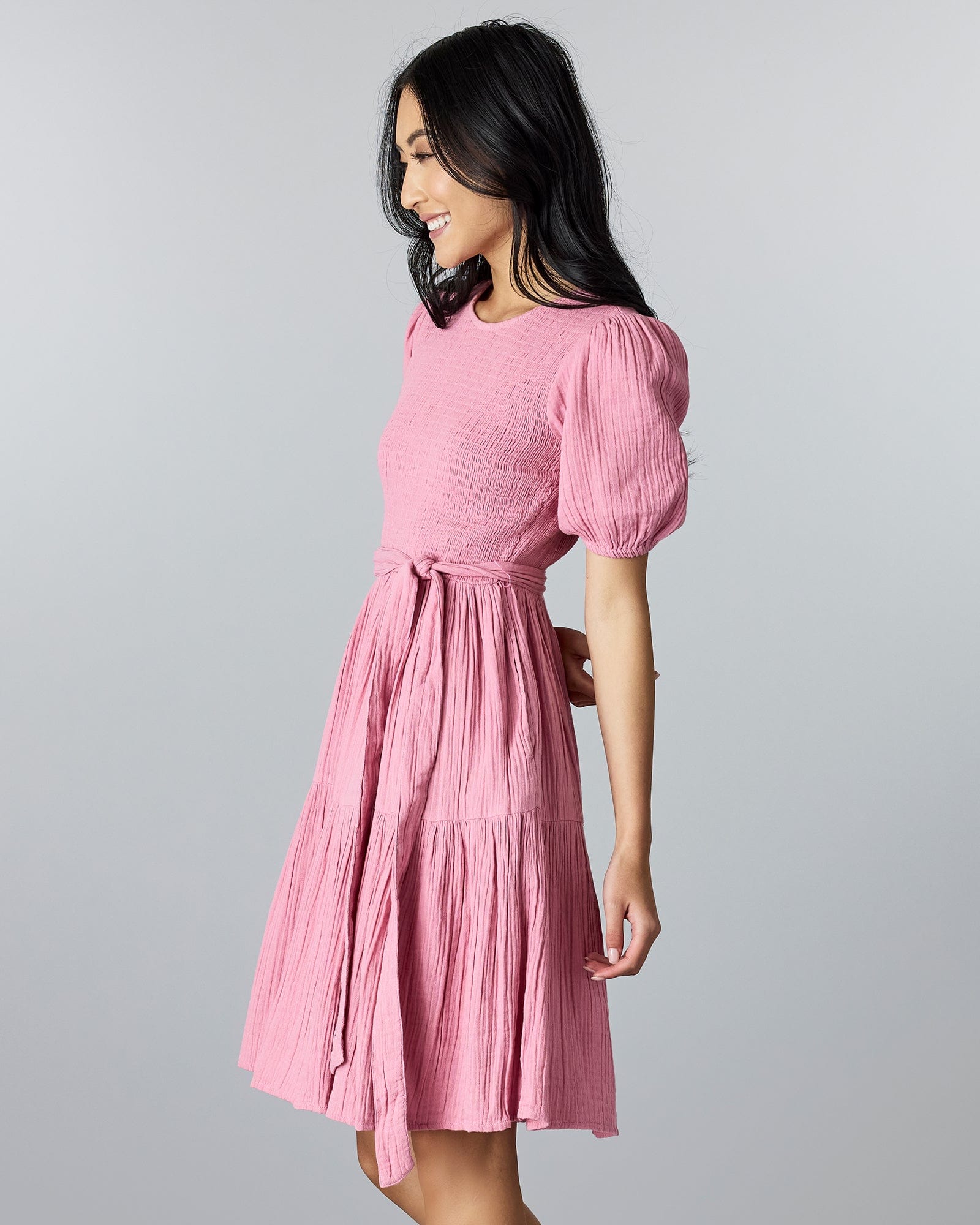Woman in a pink, short sleeved, knee-length dress with smocking on bodice and a tie at the waist.