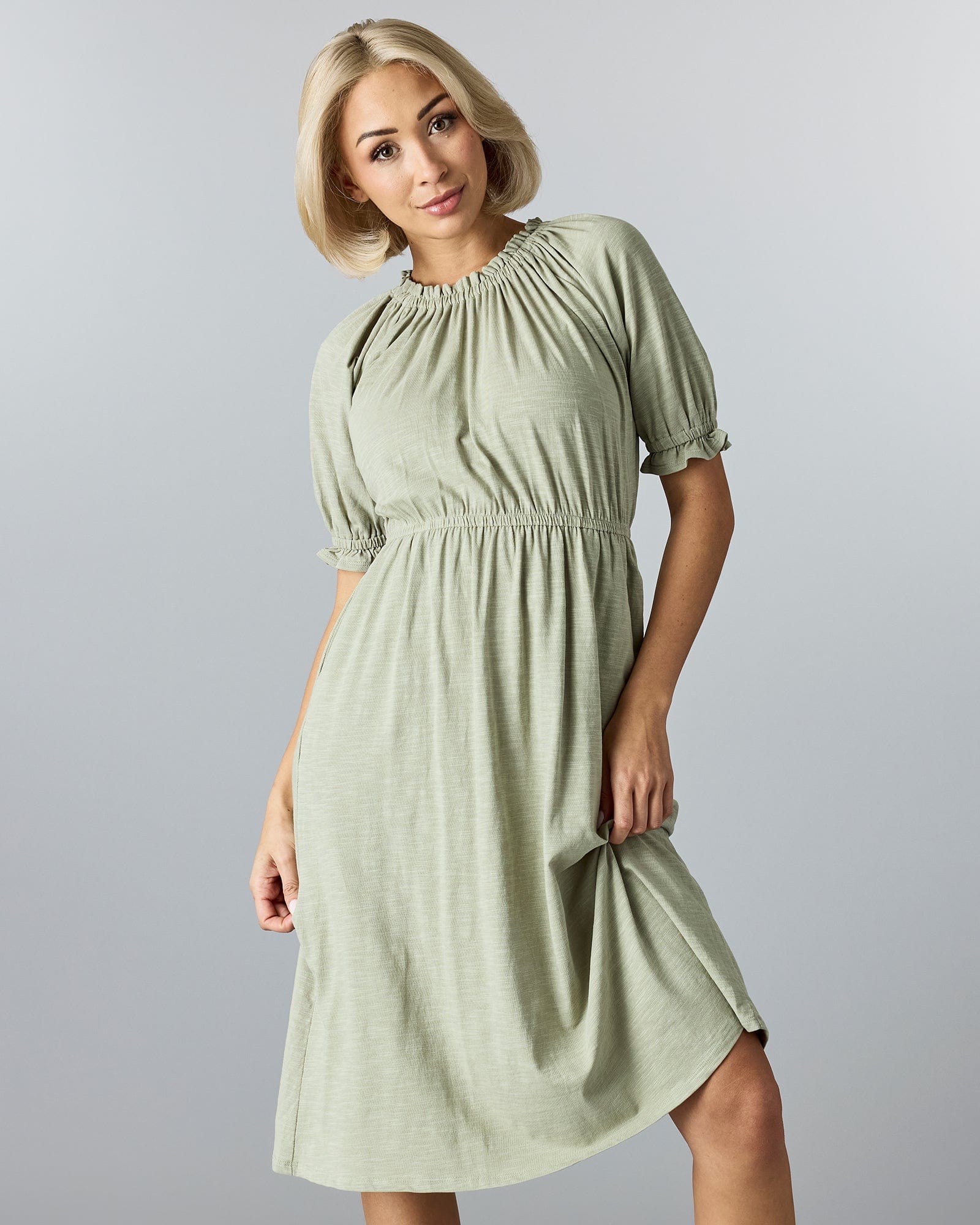 Woman in a green, knee-length, short sleeve dress with a high neckline and ruffles at neck and arms.